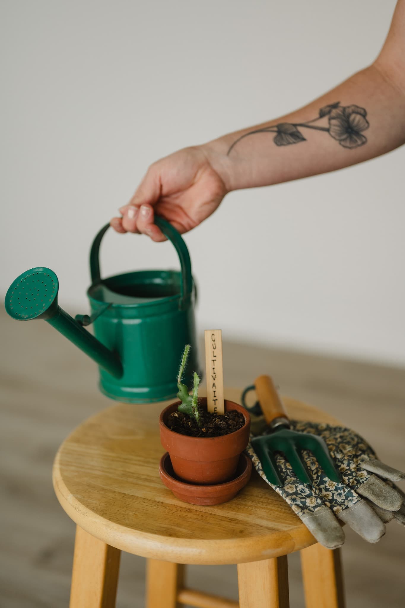 A green watering can held over Grandma Donna's clay pot, gloves, and hand fork on a wooden stool